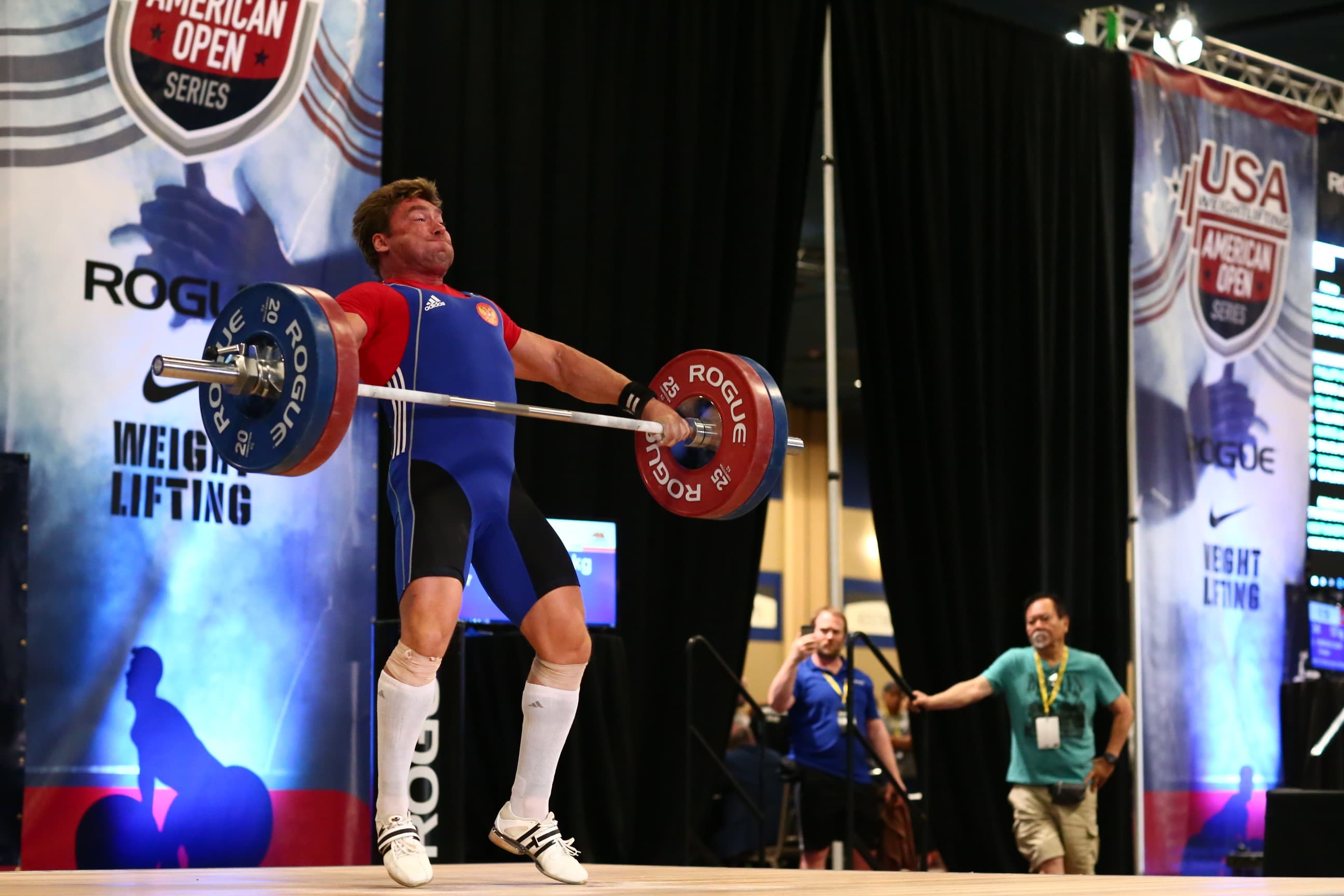 Male weightlifter in a blue singlet lifting a heavy barbell overhead at a competition.