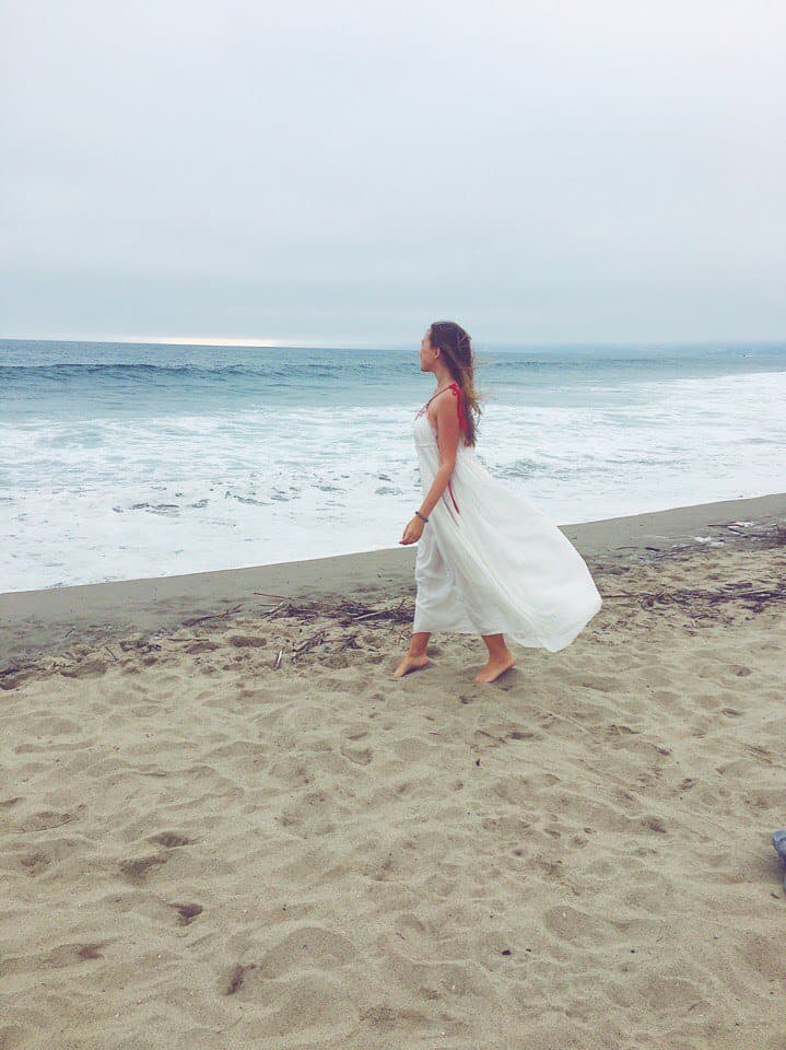 Woman in a flowing white dress walking on a sandy beach toward the ocean.