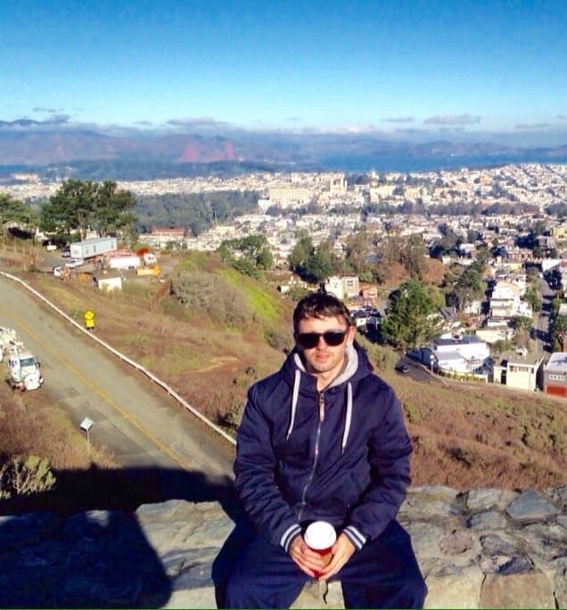Man in sunglasses and blue jacket sits on a wall overlooking a sprawling city.