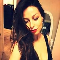 Woman with long dark wavy hair and red lipstick looking down in a black top.